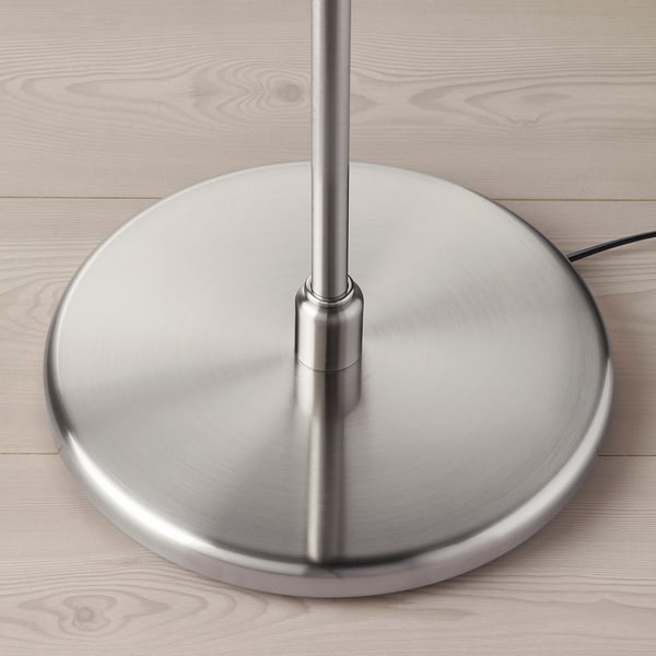 Niklowana podstawa lampy na drewnianej podłodze. Ma solidny design i metaliczne wykończenie.