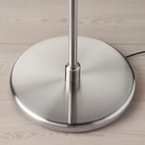 Niklowana podstawa lampy na drewnianej podłodze. Ma solidny design i metaliczne wykończenie.