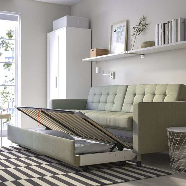 Nowoczesna, szara rozkładana sofa otwiera się do łóżka, mieści się pod minimalistyczną półką, otoczoną dywanem w paski.