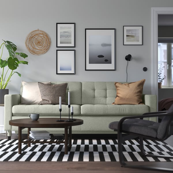 Nowoczesny salon z zieloną sofą LANDSKRONA, czarnym stolikiem kawowym, dywanem w paski, obrazami na ścianie i roślinami.