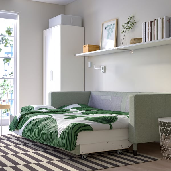 Minimalistyczna sypialnia z zieloną tropikalną narzutą na szarej rozkładanej sofie przy oknie, białymi półkami i dywanem w paski.