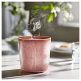 Różowawa świeca zapachowa LUGNARE, wydzielająca dym, postawiona na stole. Otoczony rozmytą zielenią. Ceramiczne, cylindryczne, o szkliwionym wykończeniu.