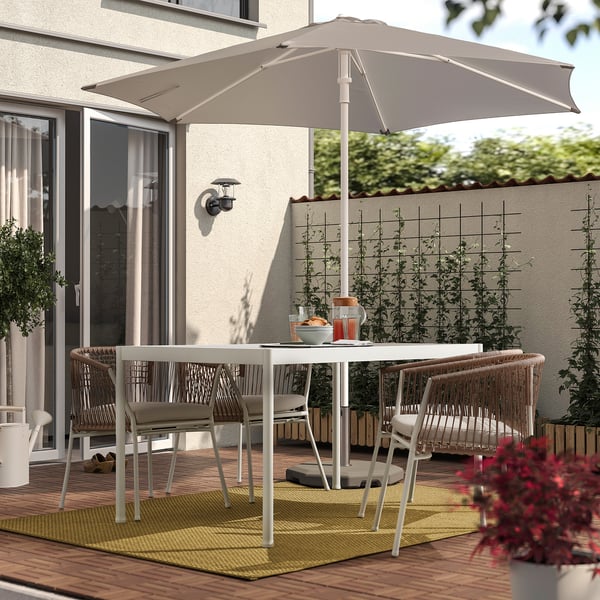 Patio na świeżym powietrzu z białym dywanem MORUM, wiklinowymi krzesłami, białym stołem jadalnym i dużym beżowym parasolem na drewnianym pokładzie, roślina na pierwszym planie.