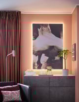 Nowoczesny salon z brzoskwiniowymi ścianami, czarną szafką, abstrakcyjną grafiką, czarną sofą, poduszkami, popcornem, rośliną, brązowymi zasłonami, światłami led.