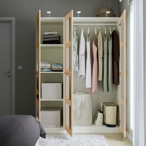 Otwarta szafa o nazwie PAX z wykończeniem z jasnego drewna, eksponująca schludnie uporządkowane ubrania i schowki. Stoi wysoko w pobliżu szarych drzwi i łóżka.