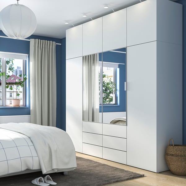 Nowoczesna sypialnia z białym schowkiem PLATSA, lustrzanymi drzwiami, łóżkiem w kratkę, szarymi zasłonami, wiklinowym koszem.