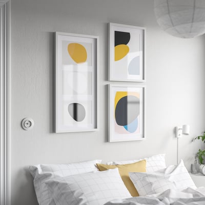 Trzy kolorowe oprawione dzieła sztuki nad łóżkiem. Geometryczne kształty w kolorze żółtym, czarnym, szarym i niebieskim. Największe poduszki z białą siatką wyśrodkowaną na ramie.
