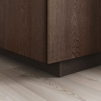 Szafka z ciemnego drewna z widocznym usłojeniem, obrzeżem z litego drewna dębowego na podstawie, stoi na jasnej drewnianej podłodze, prezentując elegancję i detal frontów SINARP.