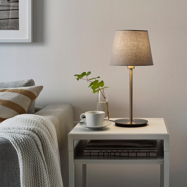 Mała, nowoczesna lampa stołowa, SKAFTET, szara, elegancki design, pasuje obok kanapy z filiżanką, wazonem i książkami.