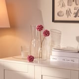 Wazon z różowymi sztucznymi kwiatami na białej komodzie, w pobliżu książek i lampy.