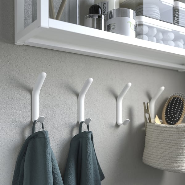 Białe podwójne haczyki na ścianie z wiszącymi ręcznikami, półka powyżej z artykułami łazienkowymi.
