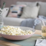 Duża miska popcornu z różowej glazurowanej ceramiki stoi na stole ze szklanką napoju. Wielkość misek porównywalna ze szkłem i dekoracjami.