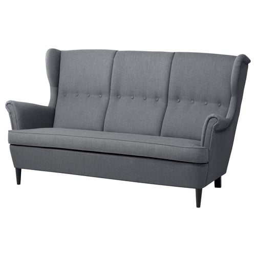 Szara 3-osobowa sofa IKEA STRANDMON z wysokimi, skrzydełkowymi oparciami, pikowanymi poduszkami i ciemnymi drewnianymi nogami.