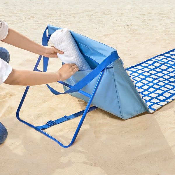 Osoba wkłada biały ręcznik do niebieskiej torby z tkaniny na leżaki STRANDÖN z zamkami błyskawicznymi na piaszczystej plaży.