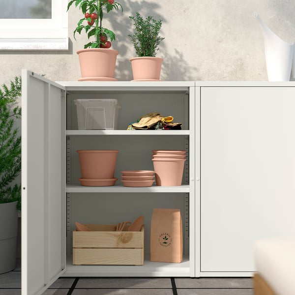 Biała szafka SUNDSÖ z regulowanymi półkami, w której znajdują się pomarańczowe doniczki z terakoty i narzędzia ogrodnicze. Idealny do przechowywania wewnątrz/na zewnątrz.