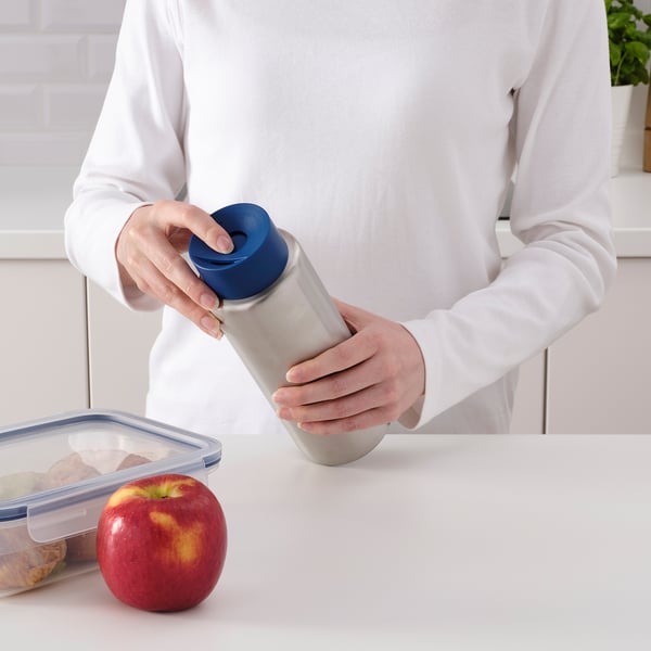 Osoba otwiera szary kubek podróżny z niebieską pokrywką, który jest odporny na rozlanie i mieści się w uchwytach samochodowych, na białym stole obok jabłka i przekąsek.