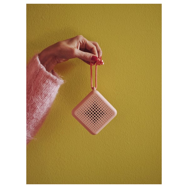 Ręka trzyma pomarańczowy, kwadratowy głośnik bluetooth VAPPEBY z małymi otworami i smyczą na tle zielonej ściany.