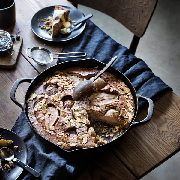 Żeliwna patelnia z ciastem na drewnianym stole. Talerz z kawałkiem ciasta, łyżką i sztućcami w pobliżu.