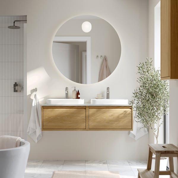 Modern bathroom with large round mirror, dual wooden vanity, white basins, and tall houseplant.