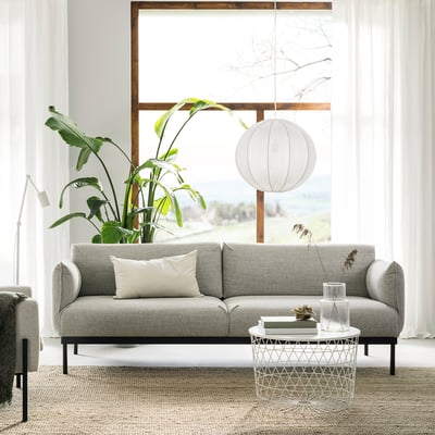 Modern living room with ÄPPLARYD gray sofa, white pillows, glass coffee table, large green plant, and hanging lamp.