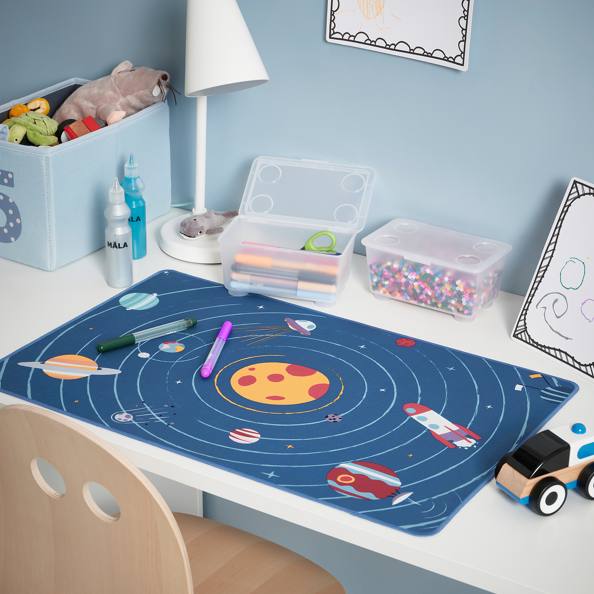 Desk with dark blue AFTONSPARV space mat, toys, lamp, and bins. Planets, spaceships, car, and water bottles nearby.