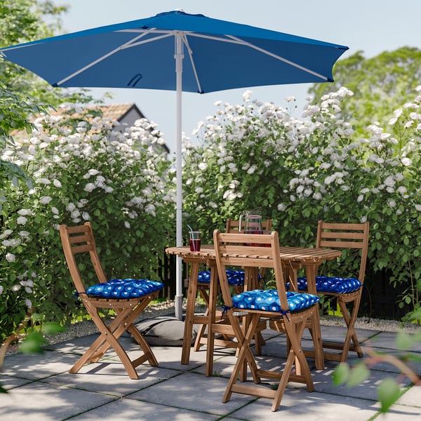 Outdoor setting with light brown wooden table and chairs, blue cushions, and matching blue umbrella, surrounded by lush white flowers.