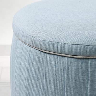 Round, light blue fabric pouffe with pleats and wooden legs.