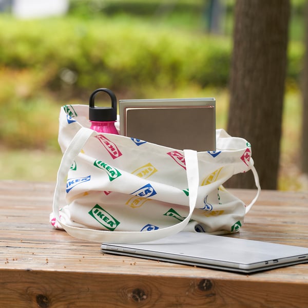 White ikea bag with colorful logos holds laptop and pink water bottle outdoors on wooden table.