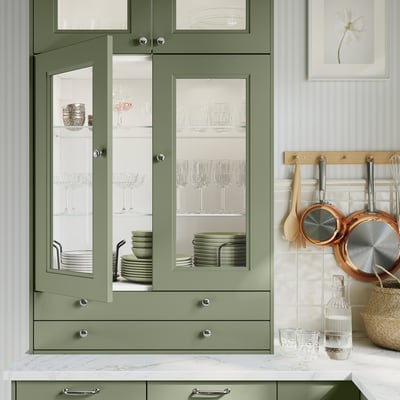 Modern kitchen with green AXSTAD cabinets, glass doors displaying dishes, marble countertop, hanging utensils, and decorative items.
