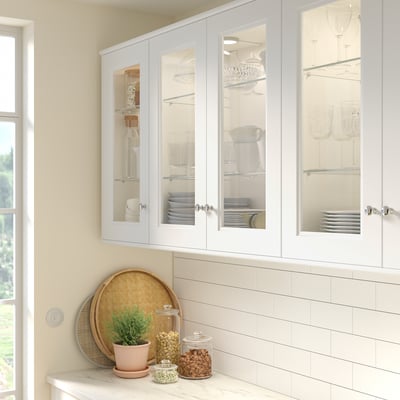 A white AXSTAD kitchen cabinet with glass doors. It holds plates, glasses, and bowls. Below, a counter holds a woven tray, plant, and glass jars.