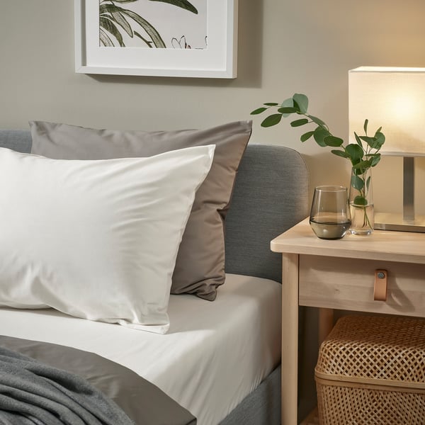 Cozy bed with white pillows, grey blanket, wicker storage, greige headboard, small nightstand holding lamp and plants.