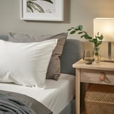 Cozy bed with white pillows, grey blanket, wicker storage, greige headboard, small nightstand holding lamp and plants.