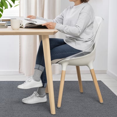 Person seated at table reading, using BALTSAR chair. Chair has transparent seat, sturdy beech legs, and bowl-shaped seat.