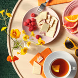 Plate with white chocolate, raspberries, and a cup of tea. Flowers and citrus slices nearby.