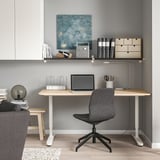 Home office setup: white desk, gray chair, laptop, wall shelf with books, art, light.