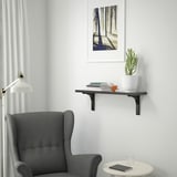 A cozy room with a gray chair and a BERGSHULT wall shelf. The shelf holds books, a plant, and a lamp. The shelf is smaller than the chair.