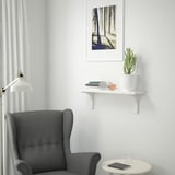 Minimalist corner with grey chair, round table, tablet, glass, wall shelf with cactus, books, and nature print.