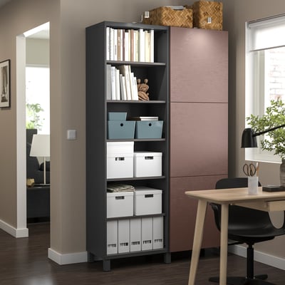 Modern office corner with BESTÅ black storage unit, wooden table, and chair. Shelves hold books and boxes. Near window with blinds.