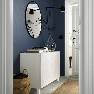 Diagonal view of modern hallway with blue wall, white BESTÅ sideboard, large mirror, basket, books, vase, and wall lamps.