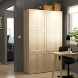 Wooden BESTÅ cabinet in office, between window & black chair. Contrasts dark walls, topped with woven boxes. Left: lamp & plant in adjacent room.