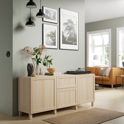 Wooden BESTÅ sideboard with 3 drawers, open shelves, sage-green wall, framed pictures, and decor on top.