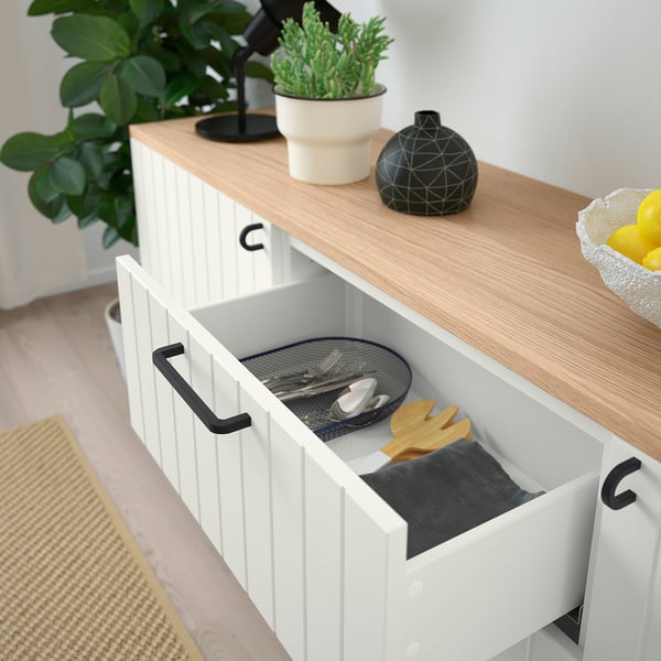 Open white cabinet with black handles, revealing plates, utensils, and cloths. Top has potted plants and black decorative vase.