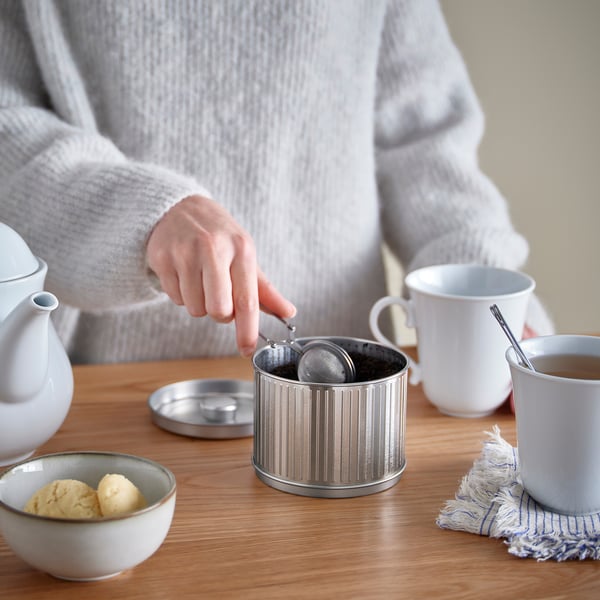 Person preparing tea at wooden table with strainer, teapot, mugs, and cookies.