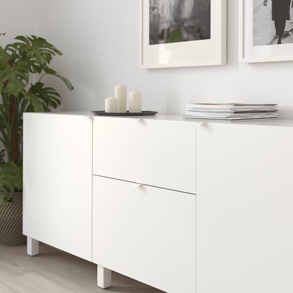 White cabinets with minimalist handles, candles, and stacked books on top, against a light wall with framed photos, and a green plant nearby.