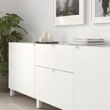 White cabinets with minimalist handles, candles, and stacked books on top, against a light wall with framed photos, and a green plant nearby.