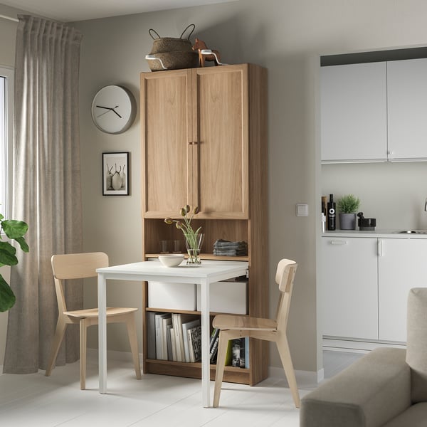 Modern dining area with light wood BILLY bookcase desk combo, white table, chairs, plant, and kitchen appliances.