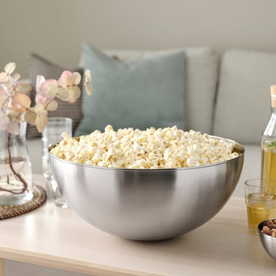 Large metal bowl filled with popcorn on a table, surrounded by glasses, a bottle, and a vase with flowers.