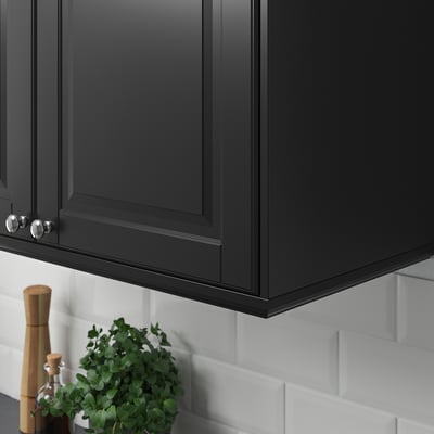 Black kitchen cabinets with silver handles and a white tiled backsplash; a potted plant and a wooden pepper grinder are placed beneath the cabinets.