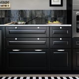 Modern black kitchen with BODBYN drawer fronts, round knobs, and white marble countertop. Dark cabinets and stainless steel appliances.