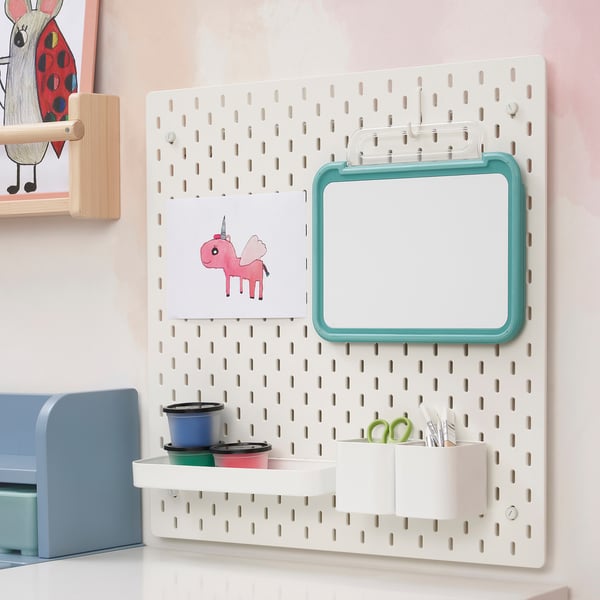 White wall-mounted pegboard with a teal-framed magnetic drawing board and containers holding colored cups and scissors.